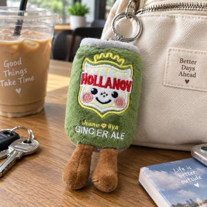 May include: A plush keyring shaped like a green soda can with a smiling face, embroidered with the text HOLLANOV and GINGER ALE, featuring two brown plush legs dangling from the bottom and a silver metal keyring attached to the top.