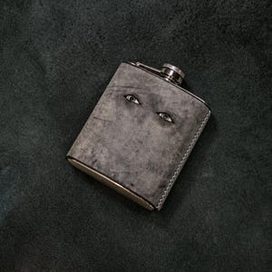 Puede incluir: Una petaca metálica rectangular envuelta en un material gris texturizado. La superficie frontal presenta dos ojos humanos realistas incrustados en el material, situados cerca de la parte superior. La petaca tiene un tapón de rosca plateado con borde estriado.