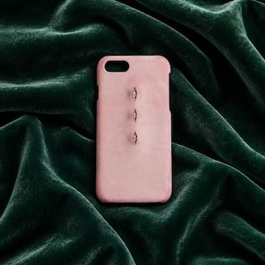 Puede incluir: Una funda de teléfono de color rosa claro con una textura suave y mate, que presenta tres pequeños adornos metálicos en forma de almendra alineados verticalmente en el centro. La funda incluye un recorte rectangular para la cámara.