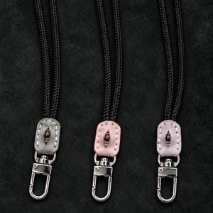 May include: Three lanyards with black braided cords and silver-toned metal lobster clasps. Each features a small, oval-shaped leather tab in gray, light pink, or lavender, stitched with white thread and centered with a small, dark, eye-shaped bead.