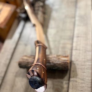 May include: A curved blade with a dark brown wooden handle featuring copper accents and small brass pins. The pommel is capped with a dark, textured circular stone inlay. The handle is shaped with a slight ergonomic curve and metallic trim.