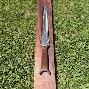 Puede incluir: Una daga de doble filo con una hoja de metal pulido en tono plateado y un mango envuelto en ante marrón con un pomo acampanado, apoyada en el centro sobre una pieza larga y rectangular de madera marrón oscuro con veta.