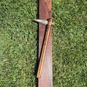 May include: A tomahawk with a metallic, pointed blade and a long, cylindrical wooden handle, featuring decorative cord wrapping near the head. The tomahawk is positioned vertically on top of a rectangular, dark brown wooden block.
