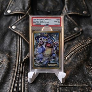 May include: A graded 2016 Japanese Pokemon Blastoise EX trading card in a clear plastic slab. The label reads 2016 POKEMON JAPANESE FA/BLASTOISE EX EXP 20th ANNIV-1st ED #092 GEM MT 10. The card features a blue turtle character with cannons.