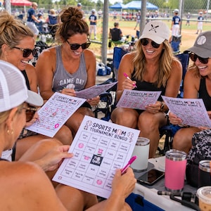 May include: Four individuals sitting in folding chairs at a baseball field, each holding a white paper game card titled Sports Mom Tournament Bingo. The cards feature a grid with various phrases and baseball icons. They are using pink pens to mark squares.