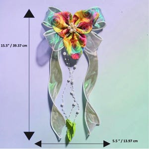 May include: A decorative bow featuring a vibrant, multi-coloured rose pattern on the top section, accented with a central cluster of white pearls. It includes long, iridescent silver ribbons and dangling pearl strands. Dimensions are 39.37 cm by 13.97 cm.