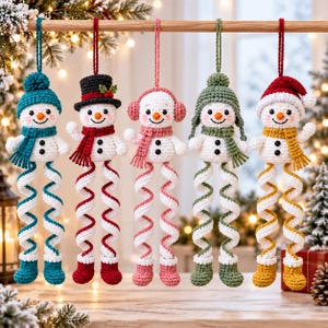 May include: Five crocheted snowman ornaments hanging from a wooden rod. Each snowman features a unique hat, scarf, and matching spiral legs with boots. The colors include teal, black and red, pink, sage green, and red and yellow accents.