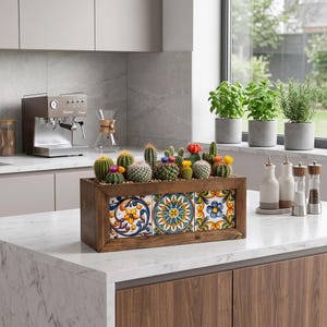 Könnte beinhalten: Ein rechteckiger Pflanzkasten aus Holz mit drei dekorativen Keramikfliesen, die farbenfrohe Blumen- und geometrische Muster aufweisen. Der Kasten ist mit einer Vielzahl kleiner grüner Kakteen bepflanzt, die teilweise rote, gelbe und blaue Blüten tragen.