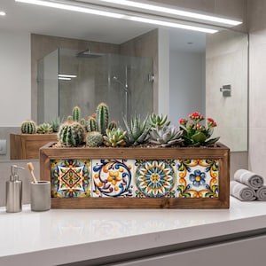 May include: A rectangular wooden planter box featuring four decorative ceramic tiles with intricate floral and geometric patterns in blue, yellow, and orange. The planter is filled with a variety of green cacti and succulents, including a flowering plant.