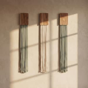 May include: Three wall hangings featuring rectangular wooden tops with long, vertical cotton cords. Two hangings have sage green cords, and the center hanging has cream-colored cords. The cords hang straight down in a uniform, parallel arrangement.