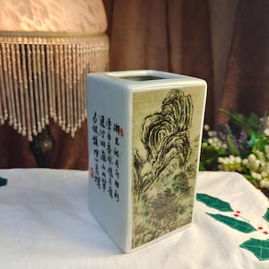 May include: A rectangular white ceramic vase featuring a green ink-wash style landscape illustration on one side and black Chinese calligraphy characters with small red stamps on the adjacent side. The vase has a smooth finish and an open top.