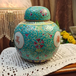 May include: A round ceramic ginger jar with a matching lid, featuring a vibrant turquoise background decorated with intricate pink floral patterns and gold-outlined scrolls. Two white circular medallions display red Chinese characters.