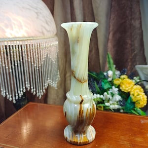 May include: A tall, slender vase crafted from polished stone with a smooth, translucent appearance. The vase features a pale, creamy green base color with prominent, swirling bands of deep brown and amber veining throughout its surface.