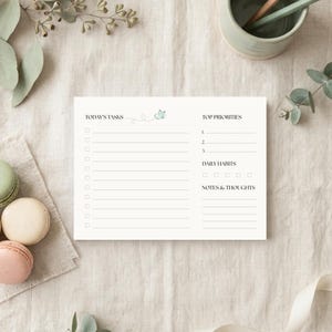 May include: A white rectangular daily planner notepad featuring sections labeled Today's Tasks, Top Priorities, Daily Habits, and Notes & Thoughts. The layout includes checkboxes and horizontal lines for writing, with a small blue floral graphic.