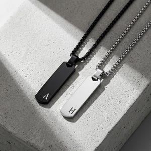 Op de afbeelding: Twee rechthoekige hangerkettingen met box-schakels. De ene hanger is matzwart met de letter A gegraveerd, en de andere is gepolijst zilver met de letter H gegraveerd. Beide hangers hebben een glad oppervlak en afgeronde randen aan de onderkant.