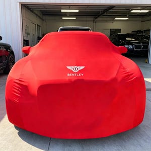 May include: A bright red car cover draped over a vehicle, featuring the white Bentley logo and the word BENTLEY printed in the centre. The fabric is smooth and form-fitting, completely obscuring the car's body while highlighting its silhouette.