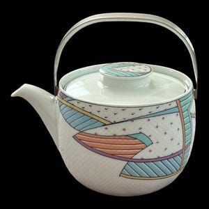 May include: A white ceramic teapot featuring a geometric pattern of light blue, orange, and purple lines and shapes over a subtle dotted texture. The teapot includes a matching lid with a small knob and a curved silver-toned metal swing handle.