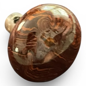 May include: A polished, rounded glass pendant featuring a swirling, marbled pattern of deep reddish-brown and translucent amber tones. The surface is smooth and reflective, with a small, clear glass loop attached to the side for suspension.