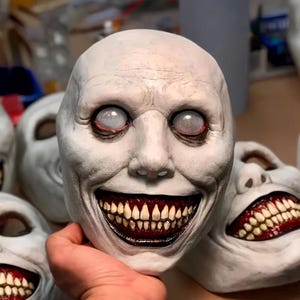 Könnte beinhalten: Eine blasse, texturierte Vollmaske mit einem breiten, zahnigen Grinsen, dunkelrotem Zahnfleisch und vergilbten Zähnen. Die Augen sind mit einem feinen weißen Netz bedeckt, und die Hautoberfläche weist Falten sowie ein mattes, cremeweißes Finish auf.