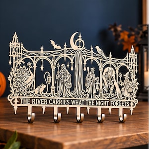 Könnte beinhalten: Ein lasergeschnittenes Wandhakenbrett aus Holz mit einem komplexen gotischen Gitterdesign, das einen Pfau, eine Ratte, eine Kapuzenfigur, einen Reisenden, ein Wesen und eine Hexe zeigt. Der Text lautet THE RIVER CARRIES WHAT THE NIGHT FORGETS.