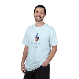 May include: A light blue short-sleeve t-shirt featuring a graphic of a baby wearing a blue and white striped party hat. Below the baby graphic, the text reads "Birthday Boy's" in a script font and "DADA" in a bold, dark sans-serif font.