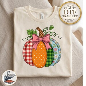 May include: A cream-colored t-shirt featuring a graphic of a pumpkin with four distinct vertical sections: red and white gingham, orange with white polka dots, green and black plaid, and blue with a floral pattern, topped with a pink bow.
