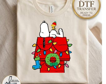 Könnte beinhalten: Ein cremefarbenes T-Shirt mit einer Grafik von Snoopy und Woodstock auf einer roten Hundehütte, die mit bunten Weihnachtslichtern, einem grünen Kranz und einem blauen Strumpf verziert ist. Die Grafik enthält den Text High Quality DTF Transfer Ready to Press.