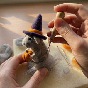 Puede incluir: Una figura de ratón de lana fieltrada gris que lleva un sombrero de bruja morado con una banda naranja. El ratón sostiene una pequeña calabaza naranja. Una aguja de fieltrar con mango de madera está cerca de la espalda del ratón.