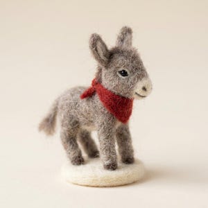 Puede incluir: Una figura de burro de lana fieltrada gris con un acabado texturizado, de pie sobre una pequeña base circular blanca. El burro presenta orejas largas y erguidas, ojos oscuros y un pañuelo triangular rojo brillante en el cuello.