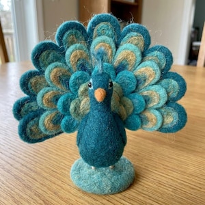 May include: A needle-felted peacock figurine featuring a vibrant teal body and a wide, fanned tail composed of layered, circular felt petals in shades of teal, light blue, and tan. The bird has a small orange beak and two dark eyes.