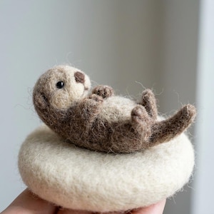 May include: A needle-felted sculpture of a small sea otter lying on its back, crafted from textured brown and cream wool fibers. The otter has a dark brown nose, small black bead eyes, and is resting on a round, cream-colored felt base.