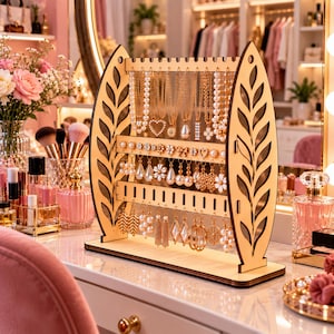 May include: A light-colored wooden jewelry display stand featuring two side panels with laser-cut leaf patterns. The stand has multiple horizontal bars with notches for hanging necklaces, earrings, and various pieces of jewelry in an organized arrangement.