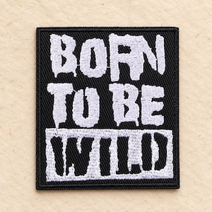 May include: A rectangular embroidered patch with a black background and white stitched text. The top portion features the words BORN TO BE in a distressed, dripping font style, while the bottom portion displays the word WILD in bold black letters.