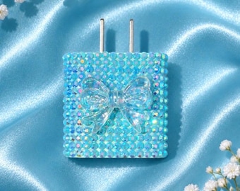 Pode incluir: Um adaptador de carregador de parede quadrado coberto com strass azul-claro iridescentes. O centro do carregador apresenta um laço decorativo de plástico transparente. Dois pinos de metal prateado estendem-se da borda superior.