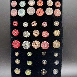 May include: A collection of assorted circular buttons arranged on a black display board. The buttons vary in size, color, and texture, featuring shades of red, green, brown, and grey, with some displaying intricate geometric patterns and four-hole designs.