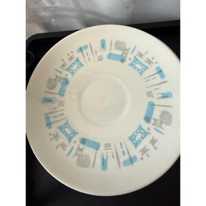 May include: A round white saucer featuring a repeating pattern of light blue and grey geometric shapes arranged in a circular band around the center. The design includes small rectangles, dashes, and stylized cross-like motifs in a balanced layout.