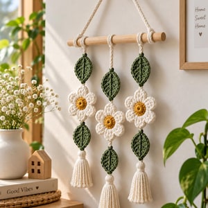 Puede incluir: Un tapiz de pared con tres hilos verticales de flores de ganchillo blancas con centros amarillos y hojas de color verde oscuro, suspendido de un listón de madera horizontal con un cordón trenzado. Cada hilo termina en una borla de hilo crema.