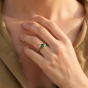 Puede incluir: Un anillo de oro con una piedra preciosa verde de corte redondo como punto focal central, flanqueada por una hilera de piedras verdes más pequeñas engastadas a lo largo de la banda. El anillo tiene un diseño abierto con extremos separados.