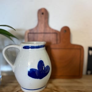 May include: A white ceramic pitcher with a blue painted floral motif, a blue band around the rim, and a blue band near the base. The pitcher features a curved handle on the left side and is positioned in front of two wooden cutting boards.