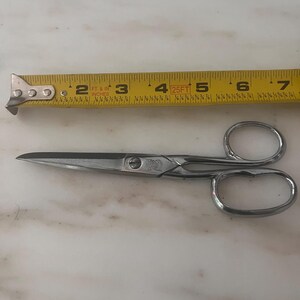 May include: A pair of silver-colored metal scissors positioned below a yellow measuring tape. The scissors measure approximately 5.5 inches in total length, with two oval finger loops and a central screw pivot point connecting the two blades.