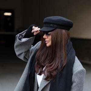 May include: A black cap with the text BY'NI embroidered on the front, paired with a long black scarf and a grey double-breasted coat. The cap features a structured crown and a short brim, while the scarf has fringed ends and a smooth texture.