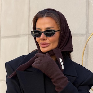 May include: A dark brown knit headscarf tied under the chin, matching dark brown knit gloves with a small white rectangular label, and black oval-shaped sunglasses. The ensemble is worn with a structured black coat featuring prominent shoulder pads.