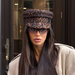 Puede incluir: Una gorra de estilo marinero con estampado de leopardo en marrón y negro, con parte superior plana, visera corta y un pequeño emblema circular negro en el lateral. La gorra se combina con gafas de media montura con marcos oscuros y lentes azul claro.