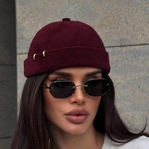 Puede incluir: Un gorro tipo docker de color burdeos intenso con un pequeño detalle metálico dorado en el lateral, combinado con gafas de sol ovaladas sin montura con cristales oscuros y un puente metálico fino, sobre una camiseta blanca de cuello redondo.