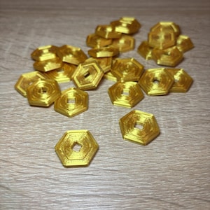 Peut inclure: Une collection de petits jetons hexagonaux de couleur or métallique, chacun doté d'une découpe carrée centrale. Les jetons présentent une surface texturée avec des lignes superposées visibles et sont dispersés dans le cadre de manière désordonnée.