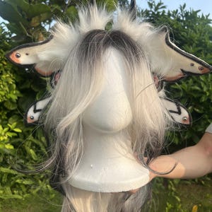 May include: A white and black synthetic wig styled with long, layered hair on a mannequin head. The wig features a decorative headband adorned with white faux fur, black pointed horns, and four wing-like appendages with black and white patterns.