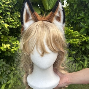May include: A pair of faux fur animal ears in shades of brown, black, and white, attached to a headband. The ears are positioned atop a white mannequin head featuring long, wavy blonde hair with bangs, styled in a layered, shoulder-length cut.