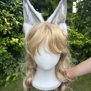 Könnte beinhalten: Ein Paar hohe, spitze Tierohren aus grau-weißem Kunstfell, befestigt an einem weißen Schaumstoff-Mannequinkopf. Der Kopf trägt langes, welliges, blondes Kunsthaar mit einem Pony, das das Gesicht umrahmt und über die Schultern fällt.