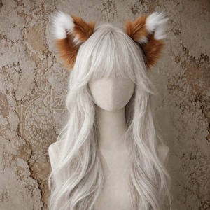 Könnte beinhalten: Ein Paar Kunstfell-Tierohren in Rotbraun und Weiß, platziert auf einem Schaufensterpuppenkopf mit langem, glattem, platinblondem Haar und Pony. Die Ohren haben eine spitze Form mit weißen Spitzen und braunen Mitten.