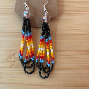 May include: Dangle earrings featuring multiple strands of small seed beads in black, light blue, red, orange, yellow, and white. Each earring hangs from a silver-toned hook and includes a single clear faceted bead at the top of the beaded fringe.